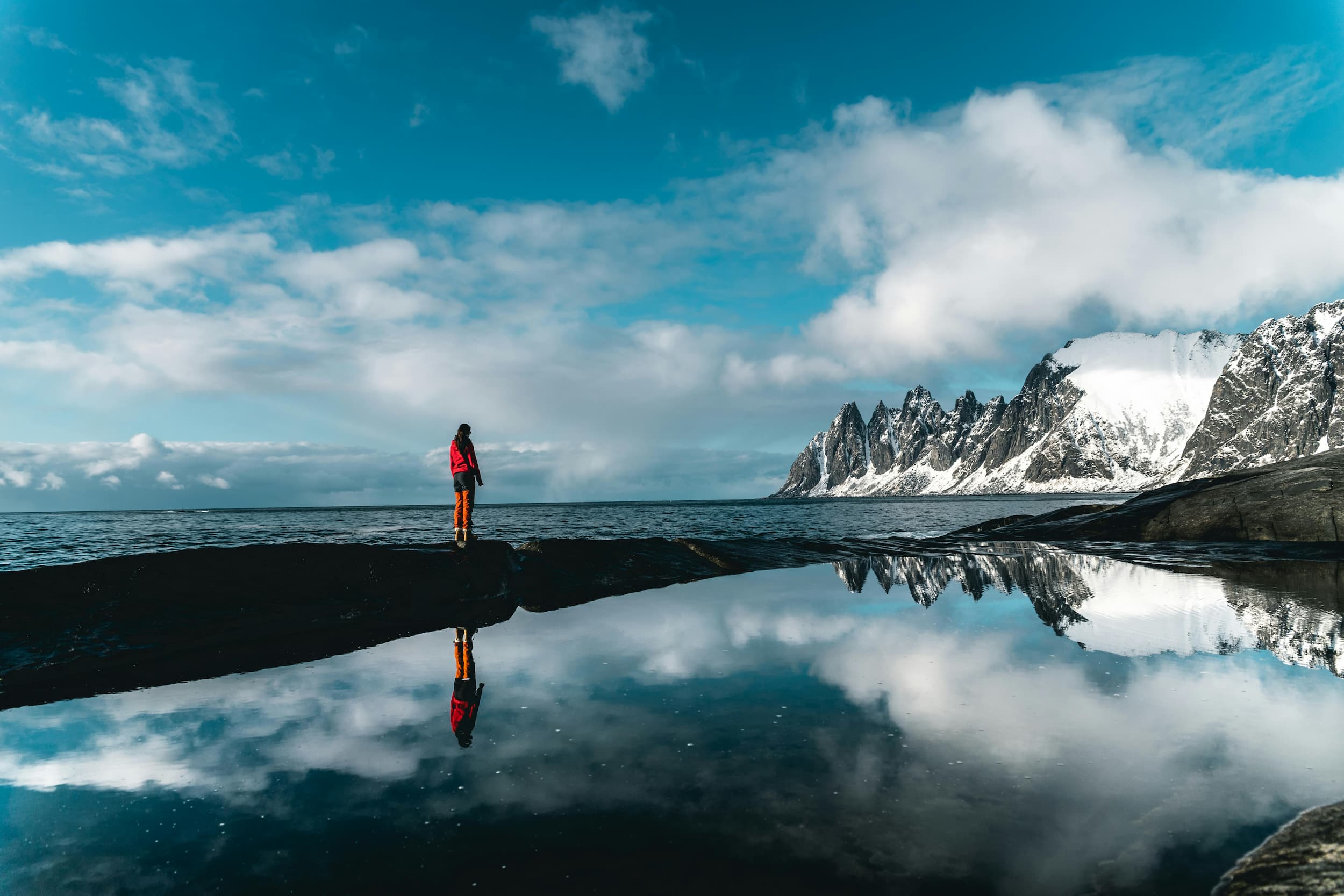 Person walking across rocks near snowy mountains and sea