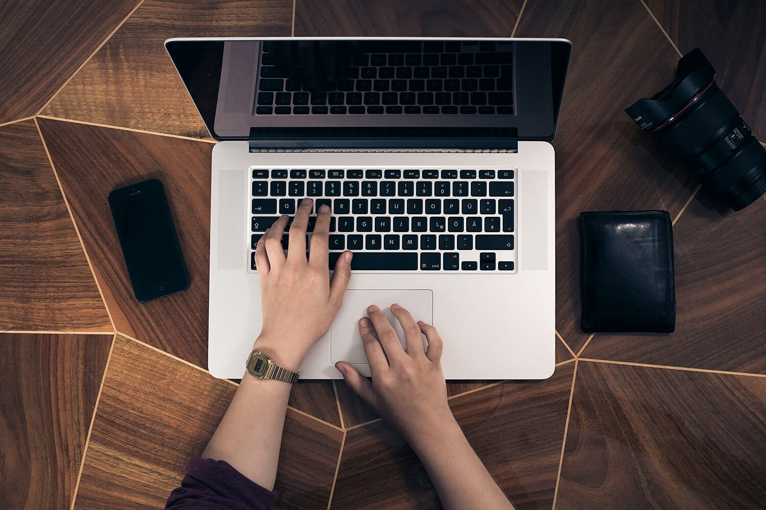 Person using a Macbook with a phone, camera lens and wallet to the sides